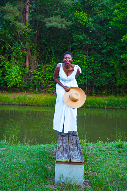Woman wearing a white dress standing on a bench near a lake holding a hat